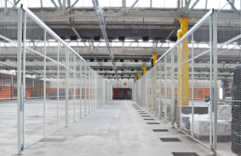 An empty industrial-style corridor with metal fencing on both sides, concrete floor, exposed metal beams, and yellow structural columns.