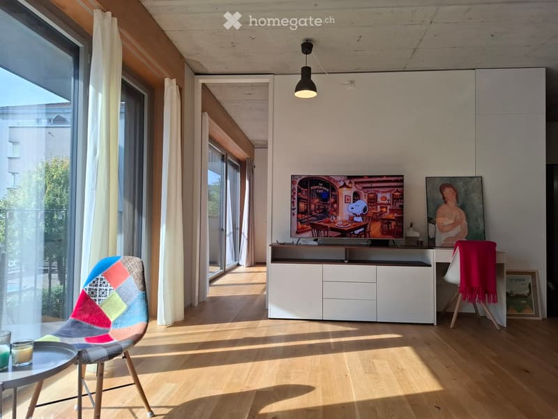 Bright living room with wooden flooring, large windows, a colorful chair, a small round table with candles, a TV on a white cabinet, and a painting on the wall.