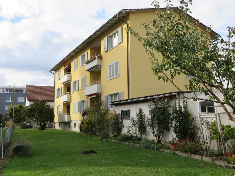 A yellow multi-story residential building with balconies, surrounded by greenery and a well-maintained lawn.