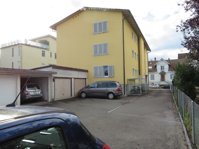 Two-story yellow building with multiple windows and shutters, attached garage with two parked cars, and a parking area with additional vehicles.