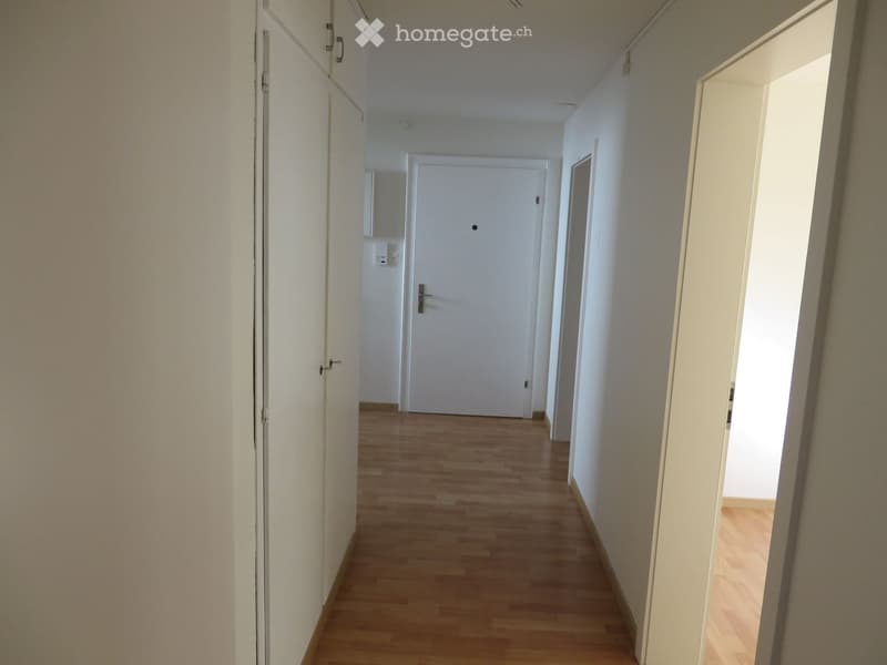 A clean, well-lit hallway with wooden flooring and white walls, featuring several doors and a small window at the end.