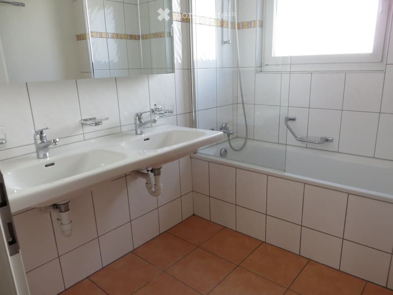 Bathroom with double sink, bathtub, and mirror. Tiled walls and wooden floor.