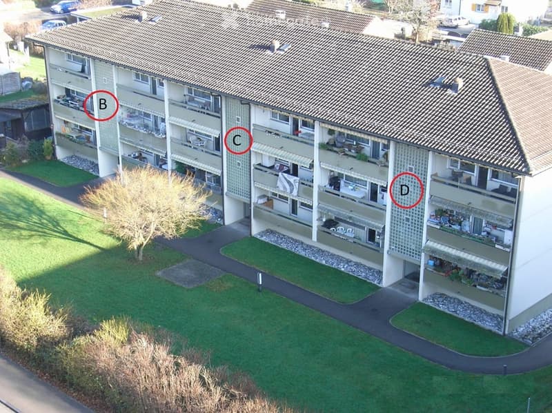 A multi-story residential building with multiple balconies and outdoor seating areas. The building features a tiled roof and is surrounded by a well-maintained lawn and paved pathways.