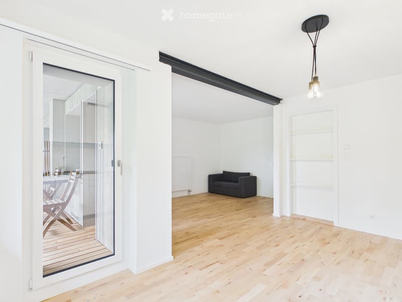Bright, open-plan living and dining area with light wooden flooring, white walls, and large glass sliding doors leading to a balcony. Features a black sofa, a dining table with chairs, and a modern hanging light fixture.