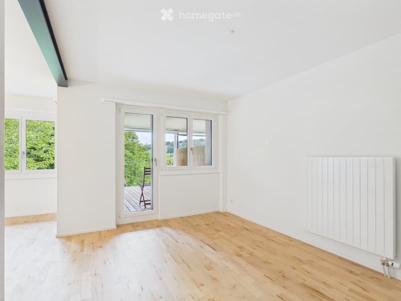 Bright, empty room with white walls, wooden flooring, large windows, and a radiator. A sliding glass door leads to a balcony with a wooden deck and a single chair visible outside.