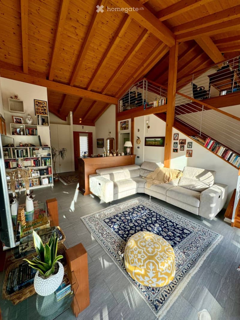 Spacious living room with exposed wooden beams, a large sectional sofa, an ottoman, a coffee table, bookshelves, a lamp, and a decorative plant.