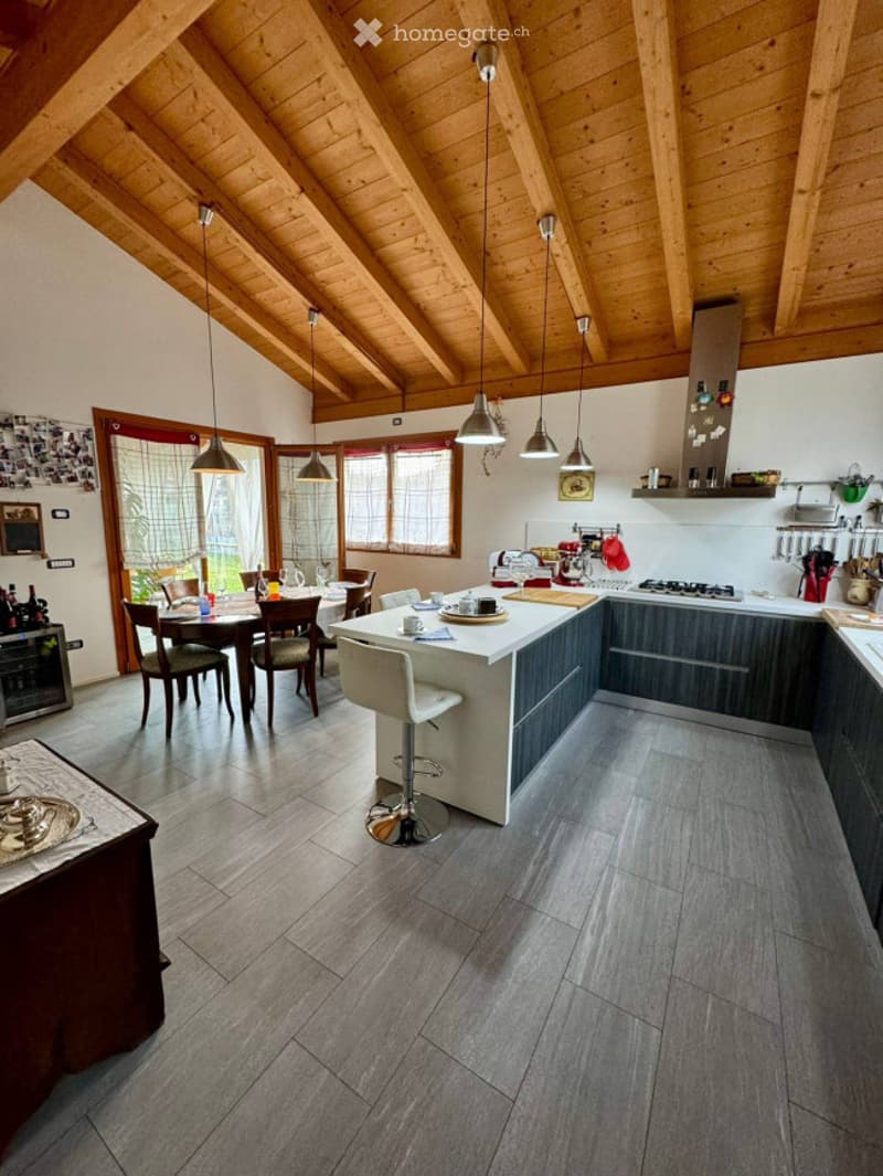 Modern kitchen with wooden ceiling beams, grey tiled floor, island with bar stool, dining area with table and chairs, and various kitchen appliances.