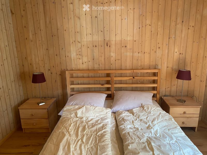 Cozy bedroom with wooden walls and floor, featuring a double bed with white bedding, two nightstands with lamps, and a simple wooden headboard.