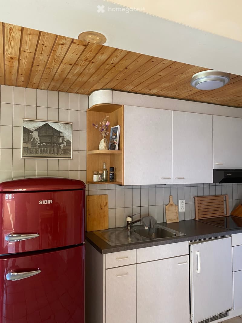 A modern kitchen with white cabinets, a red refrigerator, a sink with a faucet, and a wooden shelf holding decorative items and jars.