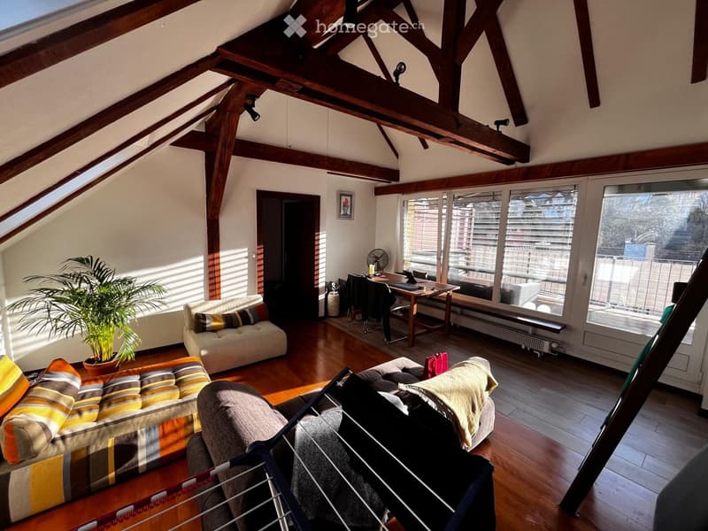 Bright attic living space with wooden beams, large windows, and a mix of seating including a sofa and armchair. Features a small desk area with a chair and a fan, along with a potted plant and a picture frame on the wall.