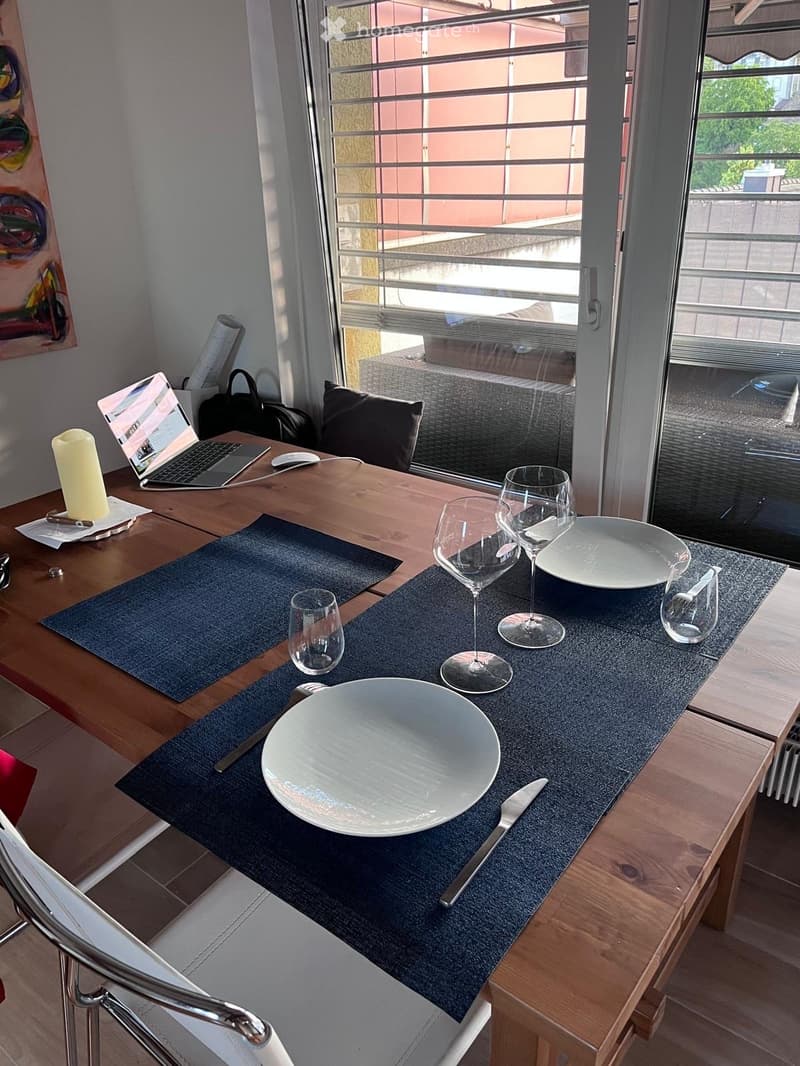 A wooden table set for two with wine glasses, plates, and cutlery, accompanied by a laptop and a candle on the table. A chair is visible to the left, and a large window with a metal railing is in the background.