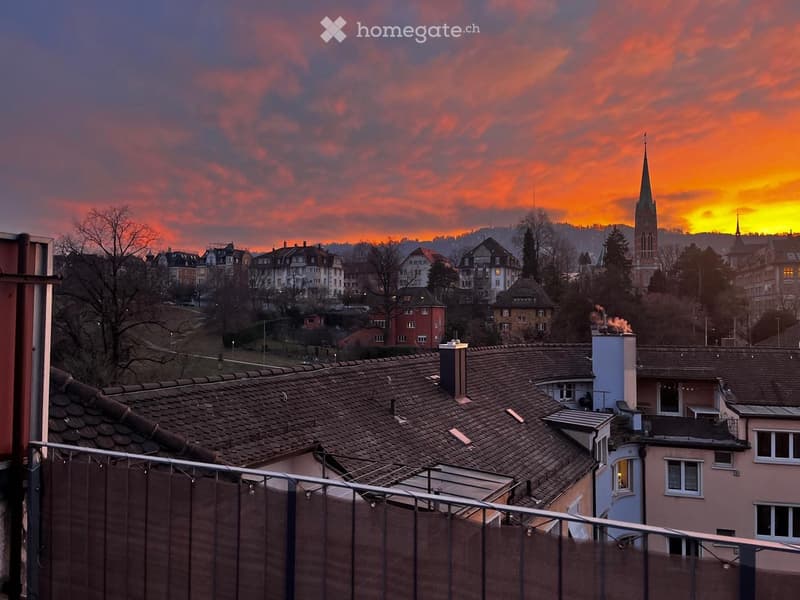 Balcony overlooking a cityscape at sunset, with rooftops, chimneys, and a distant church spire visible against a colorful sky.