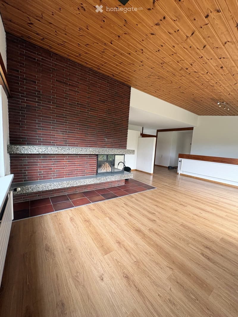 Empty room with wooden flooring, exposed wooden ceiling beams, and a brick fireplace with a glass front.