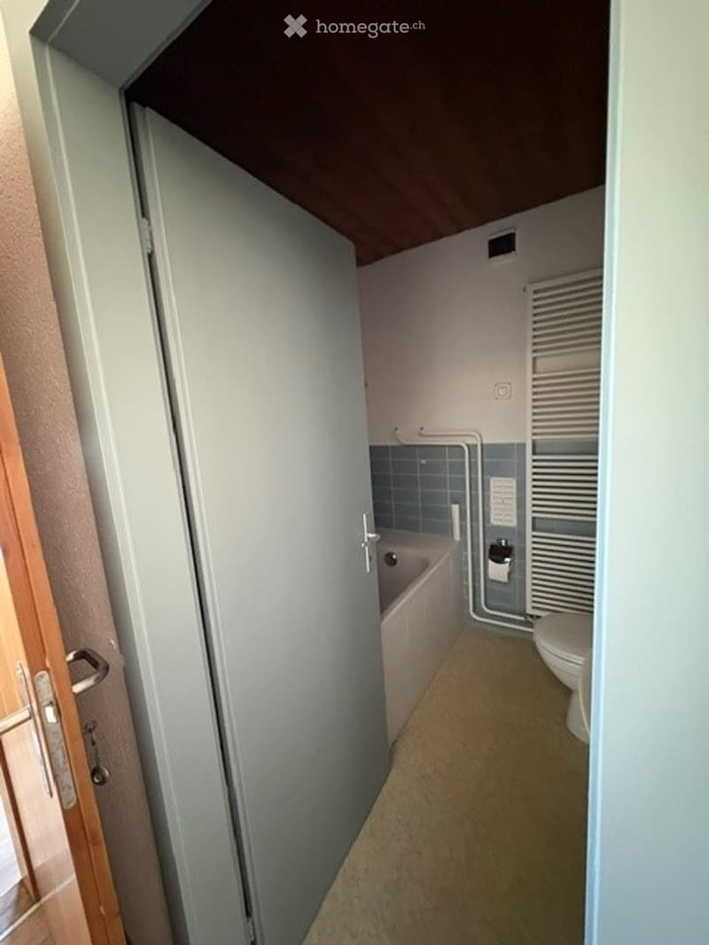 Small bathroom with a bathtub, toilet, and shower curtain. The space features light blue tiles and a wooden door frame.