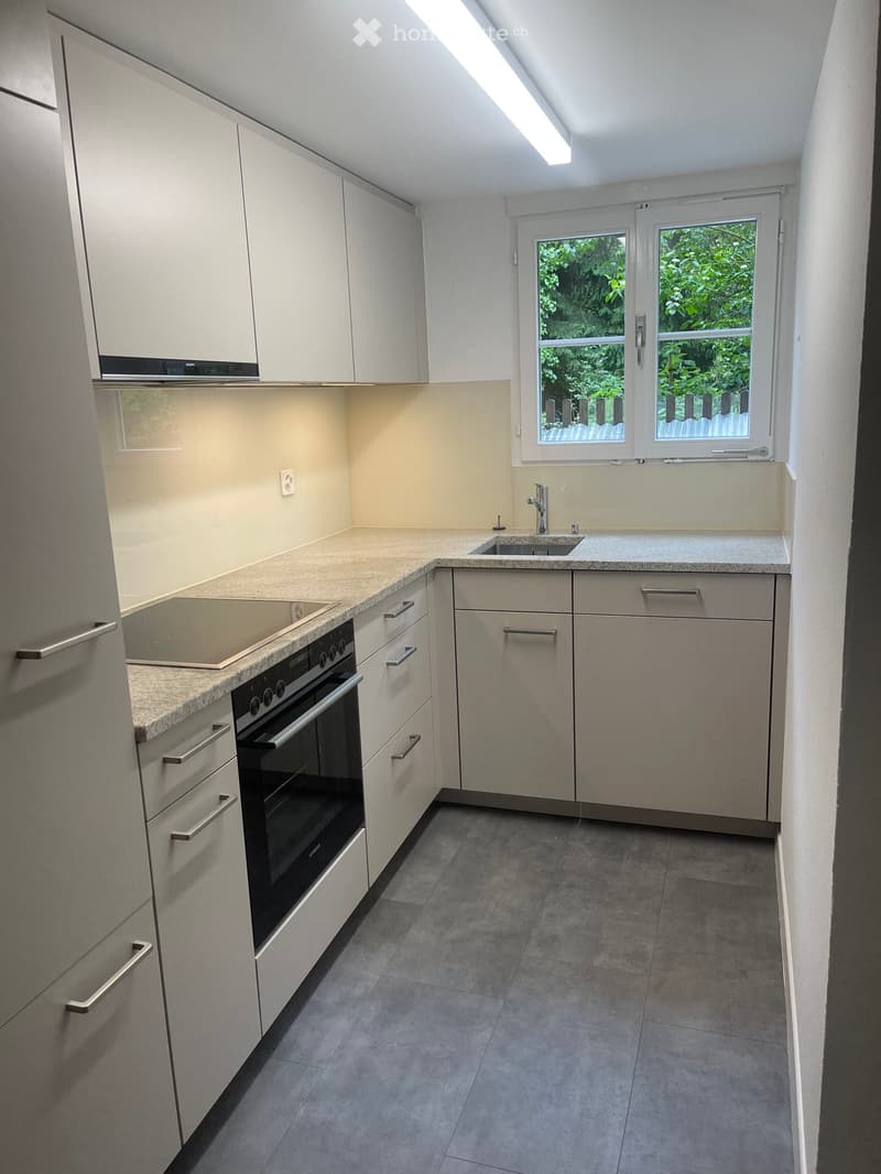 Modern kitchen with white cabinets, granite countertops, built-in oven, sink, and ample natural light from a window overlooking greenery.