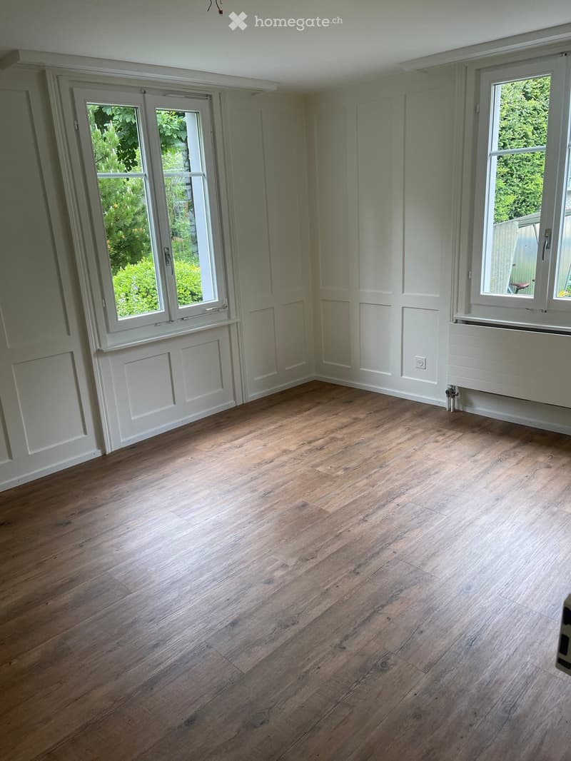 Empty room with wooden flooring, white walls, and large windows with white frames. The room has a door and a radiator visible on one wall.