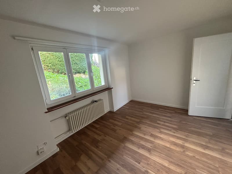 Empty room with wooden flooring, white walls, a window with a view of greenery, a radiator, and a door.