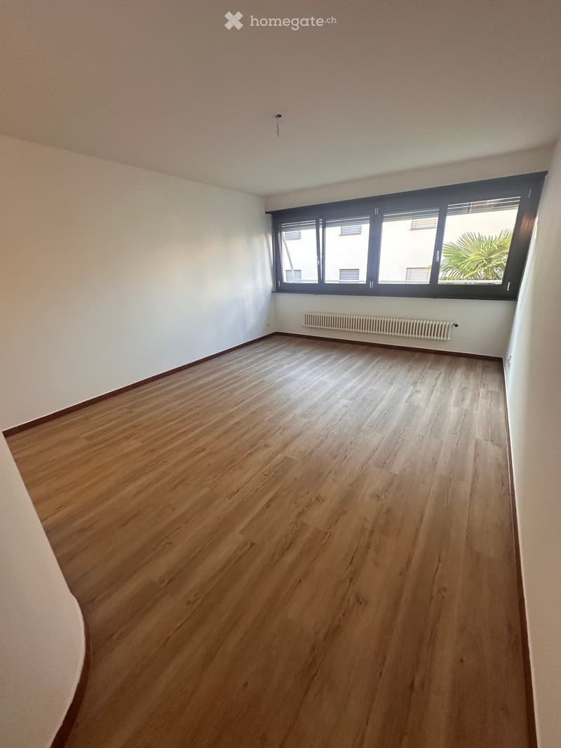 Empty room with wooden flooring, white walls, and a large window with blinds. A radiator is visible below the window.
