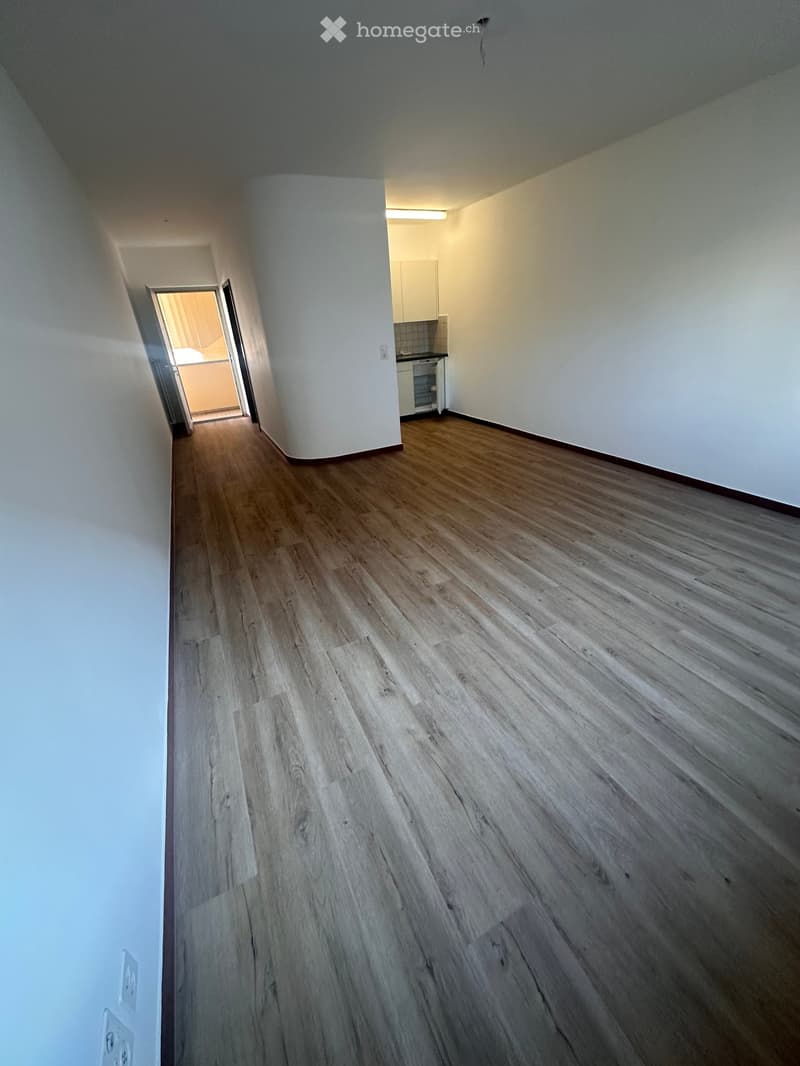 Empty hallway with wooden flooring and white walls, leading to an adjacent kitchen area.