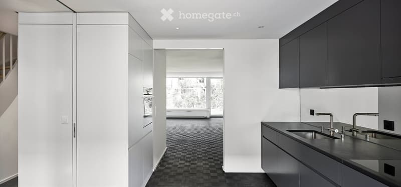 Modern kitchen with dark grey cabinets, black countertop, and stainless steel sink. Open floor plan with visible living area beyond.