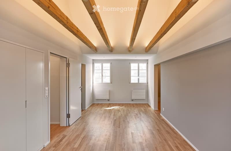Empty room with wooden flooring, exposed wooden beams on the ceiling, white walls, and two windows allowing natural light. Radiators are present on the walls.