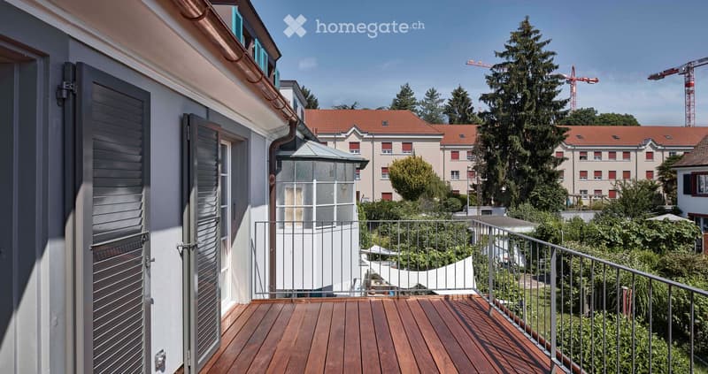 Balcony with wooden decking, metal railing, and view of surrounding buildings and greenery.