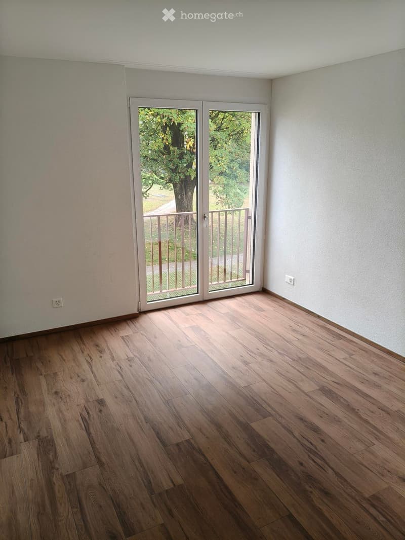 Empty room with wooden flooring, white walls, and a sliding glass door leading to a balcony with a metal railing and outdoor view.