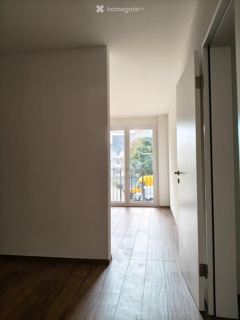 Empty hallway with wooden flooring, white walls, and a sliding glass door leading to a balcony area.