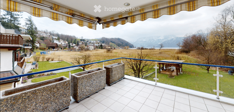 Balcony with a scenic view of a lake and mountains, featuring square planters and a striped awning.