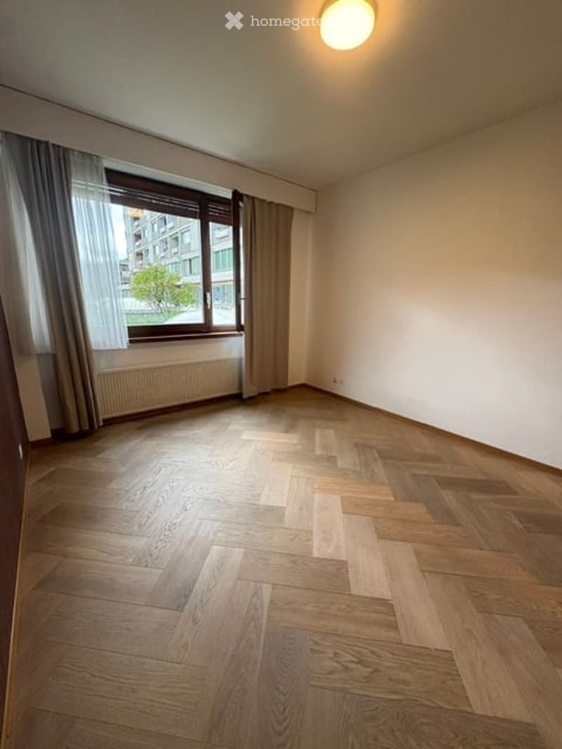 Empty room with wooden flooring, large window with curtains, radiator, and a ceiling light fixture.