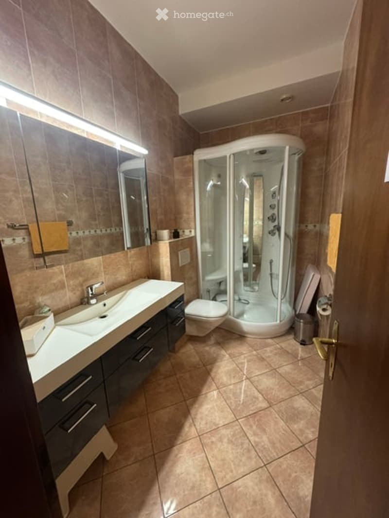 Modern bathroom with a large mirror, double sink, toilet, and a walk-in shower stall. The space features tiled walls and floor, and includes a cabinet under the sink.