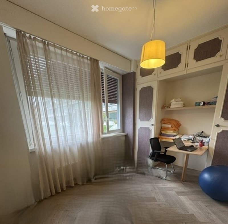 A well-lit room with wooden flooring, large windows covered by sheer curtains, a desk with a laptop, a chair, a cabinet, and a blue exercise ball on the floor.