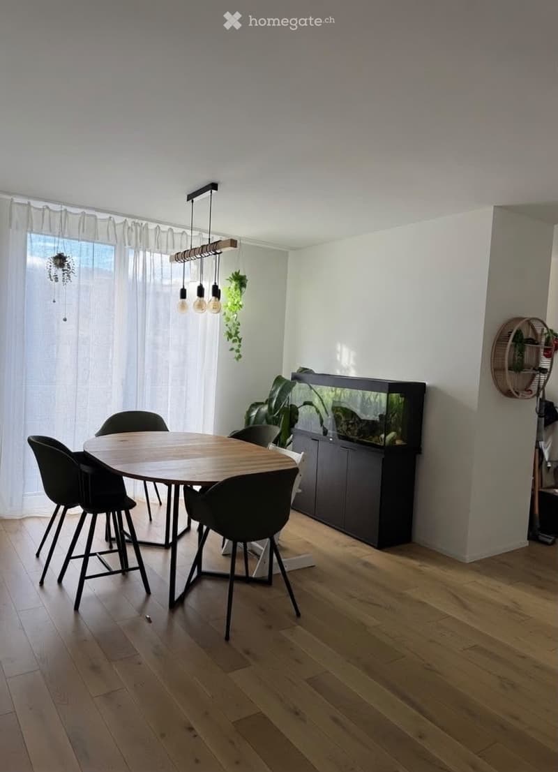 Modern dining area with a round wooden table, four black chairs, a large fish tank, pendant lights, and plants. The room features light wooden flooring and white walls.