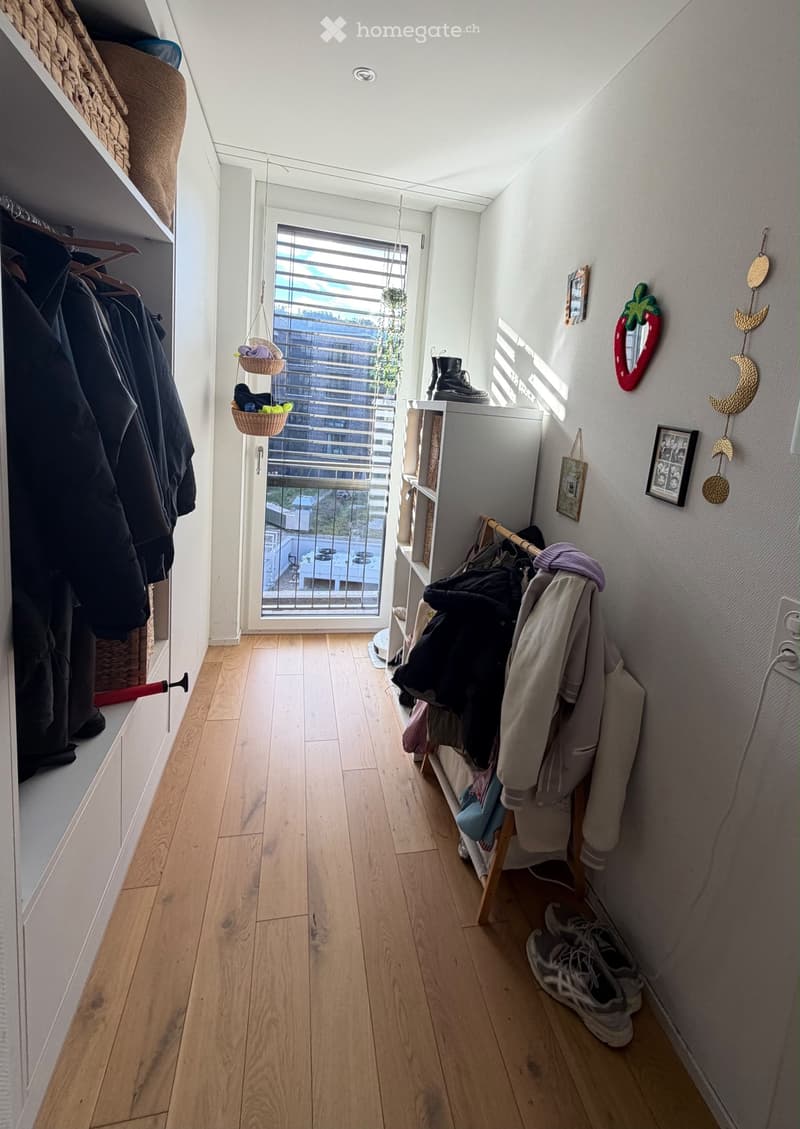 A narrow hallway with wooden flooring, white walls, and a large window with blinds. There are coats hanging on a rack, a small shelf with items, and a hanging basket with plants. The hallway leads to a balcony area with a view of the city.