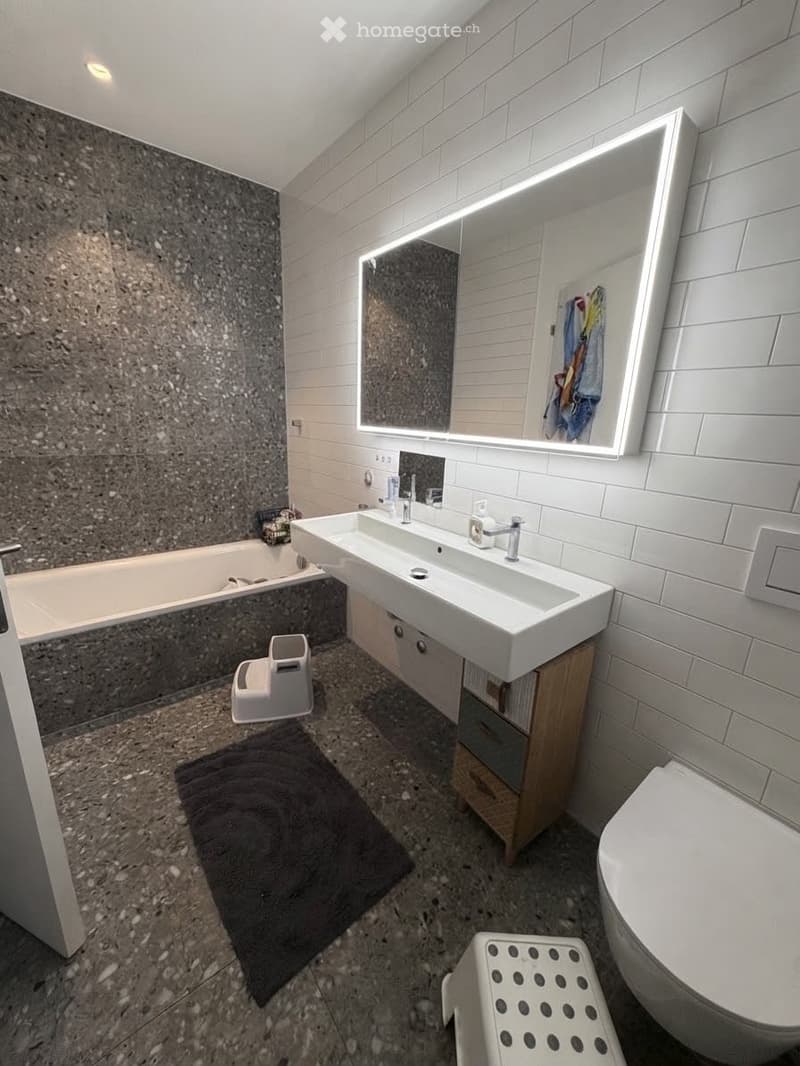 Modern bathroom with a double sink, large illuminated mirror, bathtub, toilet, and a small wooden cabinet. Includes a dark bathmat and a white stool.