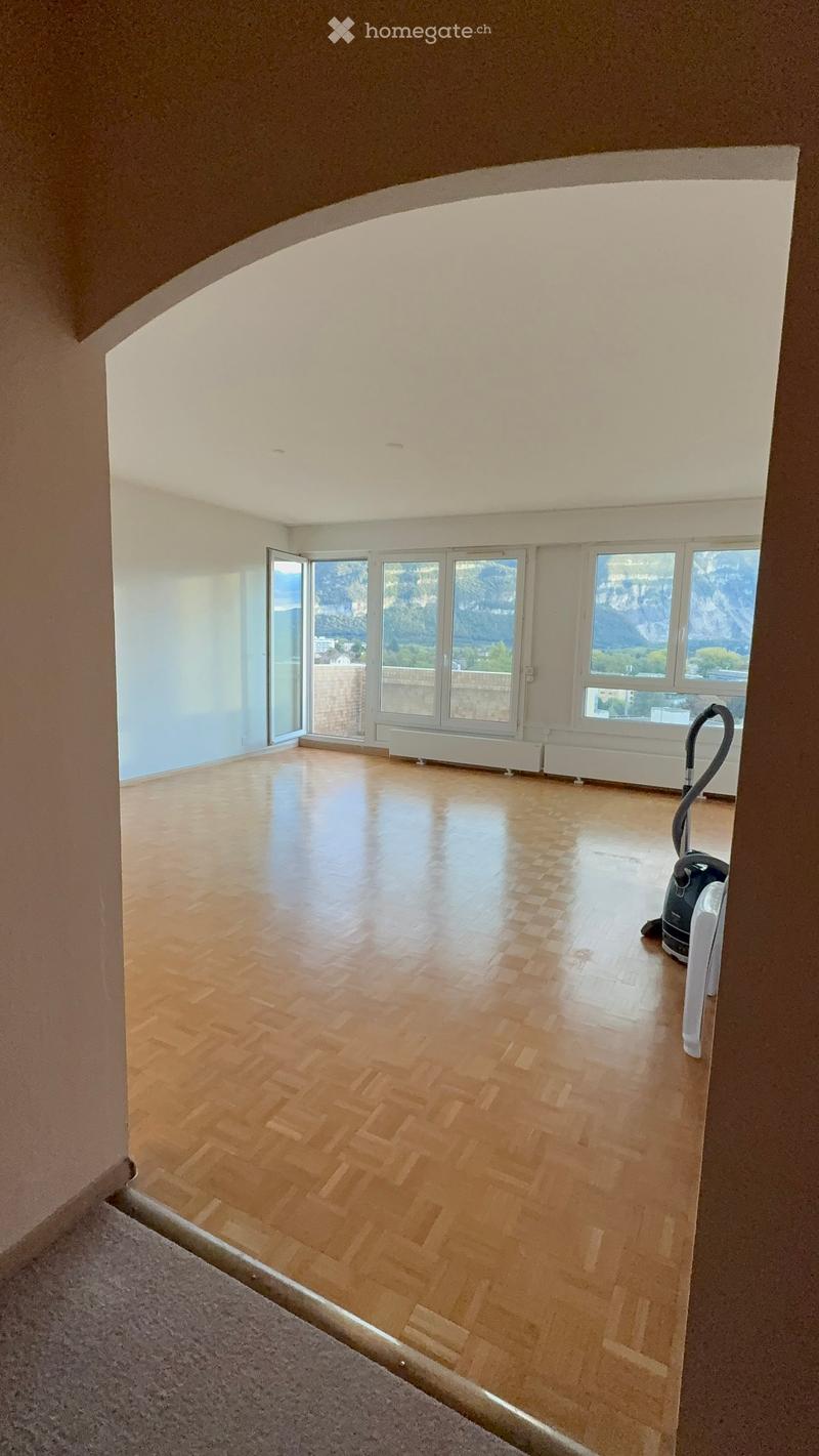 Empty room with wooden flooring, large windows offering a scenic mountain view, and a vacuum cleaner in the corner.