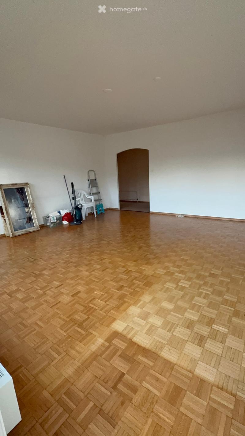 Empty room with wooden herringbone flooring, white walls, and a doorway leading to another space. Cleaning supplies and a ladder are present near the doorway.