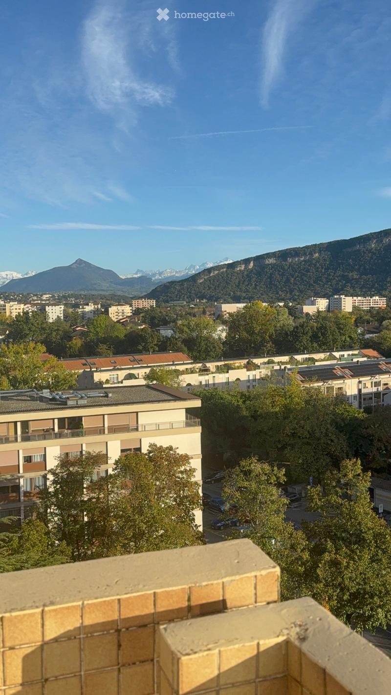 View from a balcony showing a cityscape with buildings, trees, parked cars, and distant mountains under a clear blue sky.