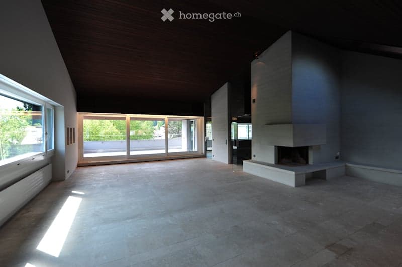 Open-concept living space with a high, slanted wooden ceiling, large windows, and a fireplace. The floor is tiled, and the room is completely unfurnished.