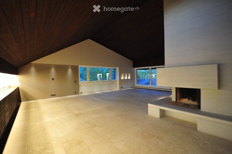 An empty room with a high, slanted wooden ceiling, large windows, and a fireplace with a built-in bench. The floor is tiled, and the walls are painted in neutral tones.