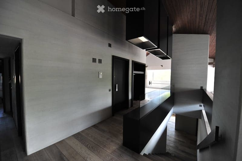 Modern hallway with wooden flooring, white walls, black doors, and a sleek black railing leading to an open area with natural light.