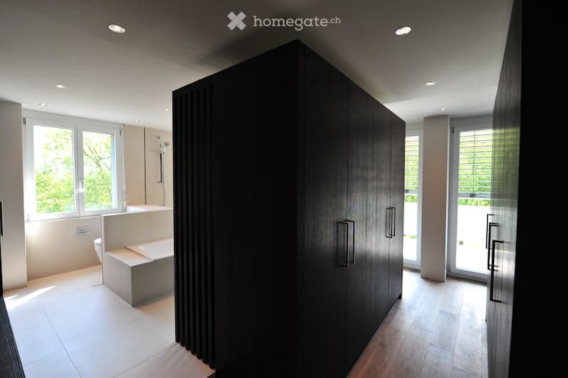 Modern bathroom with a large dark wooden cabinet, a bathtub, a toilet, and a sink. The space features large windows with blinds and recessed lighting.