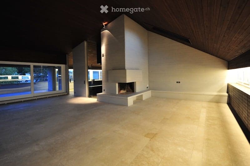 Open-concept space with a raised fireplace, large windows, and wooden ceiling beams. The floor is tiled, and the walls are white.