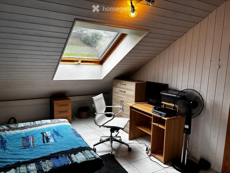 Cozy attic room with slanted wooden ceiling, skylight window, desk with computer and fan, office chair, bed with quilt, and small cabinet.