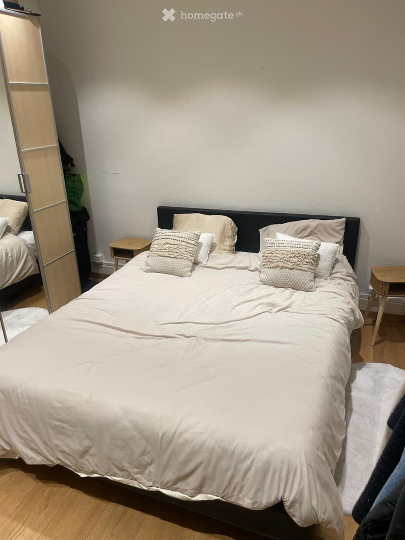 A well-lit bedroom with a large bed, nightstands, and a mirrored wardrobe. The bed is neatly made with pillows and a duvet.