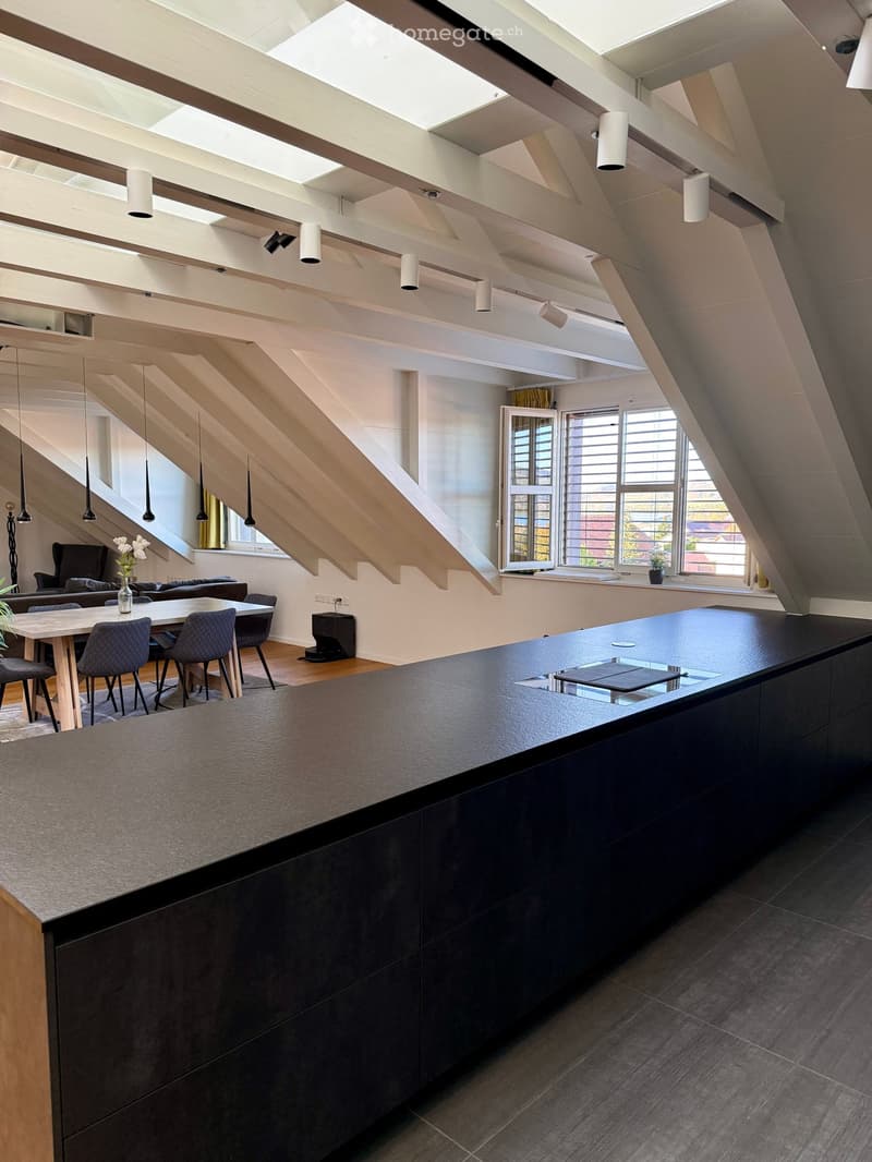 Modern open-plan living and dining area with high ceilings, exposed wooden beams, pendant lights, and large windows. Features a dark kitchen island, dining table with chairs, and a comfortable sofa area.