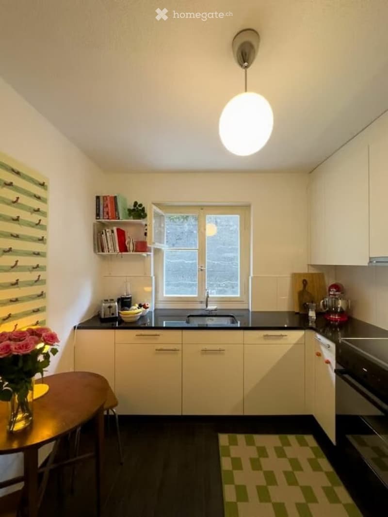 Modern kitchen with white cabinets, black countertops, a round pendant light, and a small dining area with a table and chair. Includes a window above the sink, a bookshelf with books and decorative items, and various appliances like a blender, toaster, an
