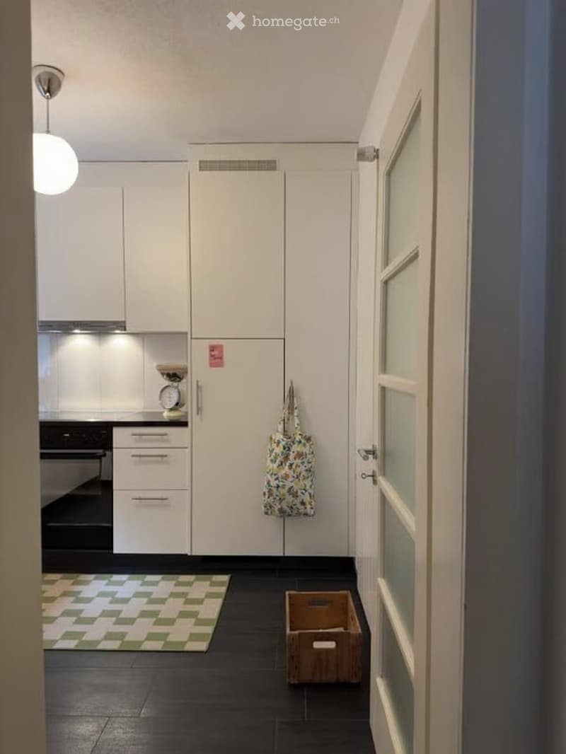 A modern kitchen with white cabinets, a black oven, and a small rug on the floor. A handbag is hanging on the wall, and a wooden box is placed near the door.