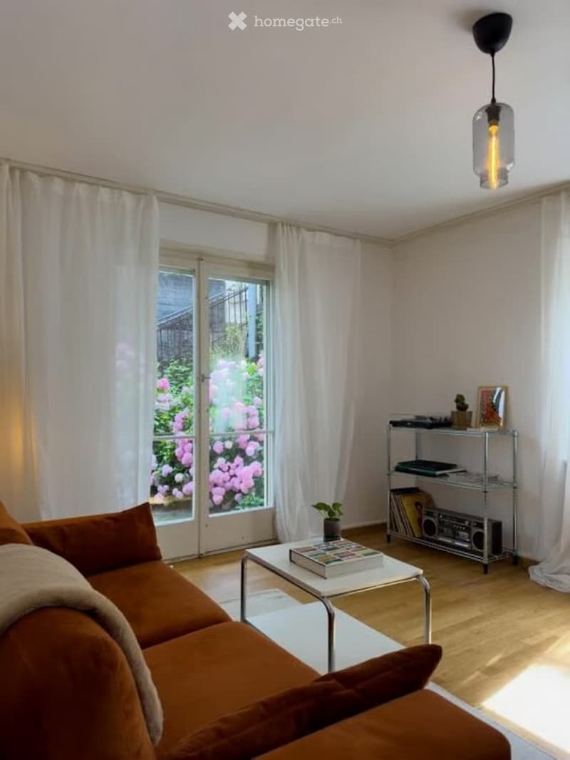 Modern living room with large windows, a brown sofa, a white coffee table, a floor lamp, a metal shelf with books and a boombox, and a small plant on the floor.