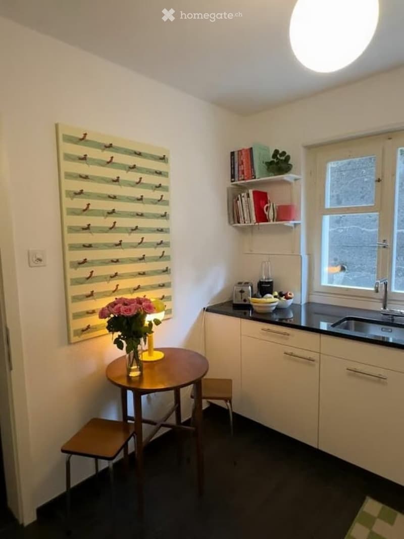 The image shows a small, modern kitchen with white cabinets, black countertops, and a round wooden dining table with two stools. A yellow lamp sits on the table, and a vase of pink flowers is placed on the table. A window with frosted glass is visible, al
